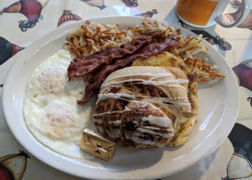 A plate of breakfast featuring eggs, bacon, hash browns, and a cinnamon roll drizzled with icing.