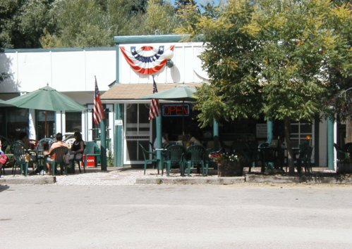 A quaint café with outdoor seating, green umbrellas, and patriotic decorations, featuring an "OPEN" sign.