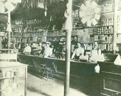 Historic grocery store interior with shelves of canned goods, a long counter, and staff members in aprons.