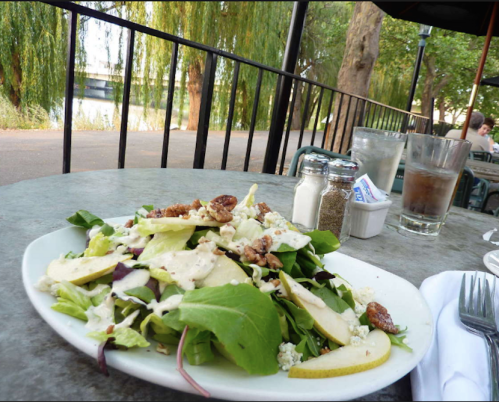 A fresh salad with greens, apples, and nuts on a table by a riverside, with a glass of water and condiments nearby.