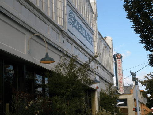 Exterior of a building with signs for "Ramblin' Jacks" and a pizza place, surrounded by greenery and a clear blue sky.
