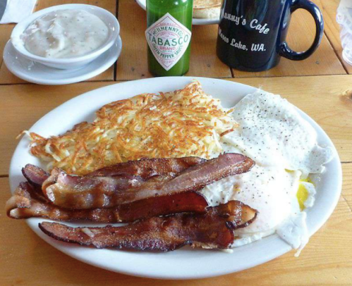 A plate of breakfast featuring crispy bacon, fried eggs, and hash browns, with a coffee mug and condiments in the background.