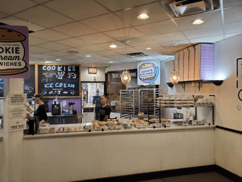 A dessert shop interior featuring a counter with cookie sandwiches, ice cream, and a staff member serving customers.