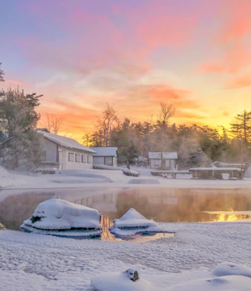 A serene winter landscape with a snowy shore, calm water, and a colorful sunrise reflecting on the surface.