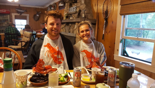 A couple wearing lobster bibs sits at a table with seafood, drinks, and a cozy cabin backdrop.