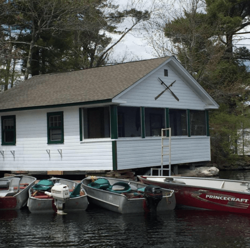 A white cabin on stilts by the water, surrounded by several boats moored nearby. Trees are visible in the background.