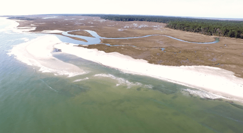 Aerial view of a sandy beach with winding waterways and lush greenery in the background.