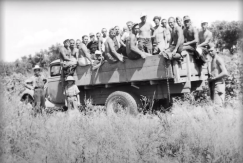 A group of shirtless men sitting on a truck in a grassy area, with a few standing nearby, in a vintage black-and-white photo.