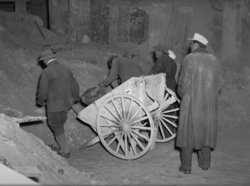 Black and white image of workers using a wheelbarrow in a construction site, surrounded by debris.