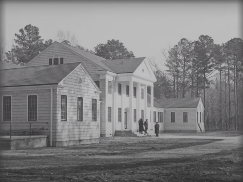Historic building with white and wooden exteriors, surrounded by trees, with people walking nearby.