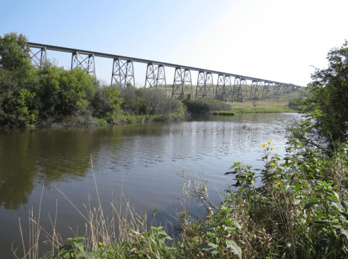 A tall railway bridge spans a calm river, surrounded by lush greenery and a clear blue sky.
