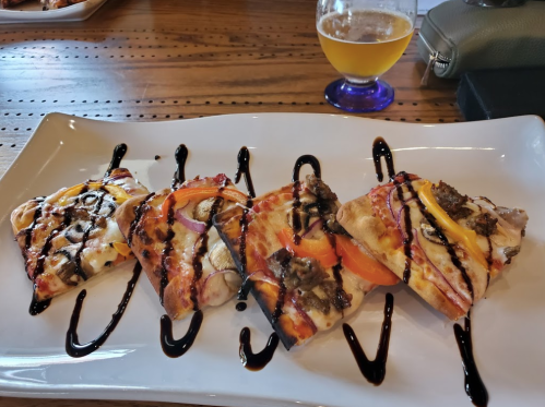 A plate of four gourmet pizzas topped with vegetables and drizzled with balsamic glaze, alongside a glass of beer.