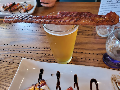 A grilled bacon strip rests on the rim of a pint glass of beer, with a plate of food in the background.