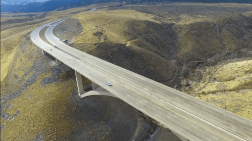 Aerial view of a highway bridge spanning a hilly landscape with sparse vegetation and winding roads.