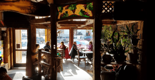 A cozy café interior with wooden beams, stained glass, and patrons enjoying drinks amidst plants and natural light.
