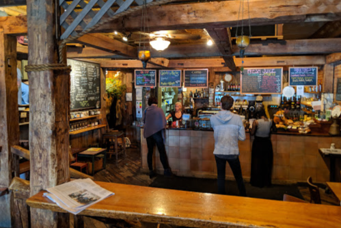 A cozy café interior with wooden beams, a chalkboard menu, and customers at the counter. Warm, inviting atmosphere.