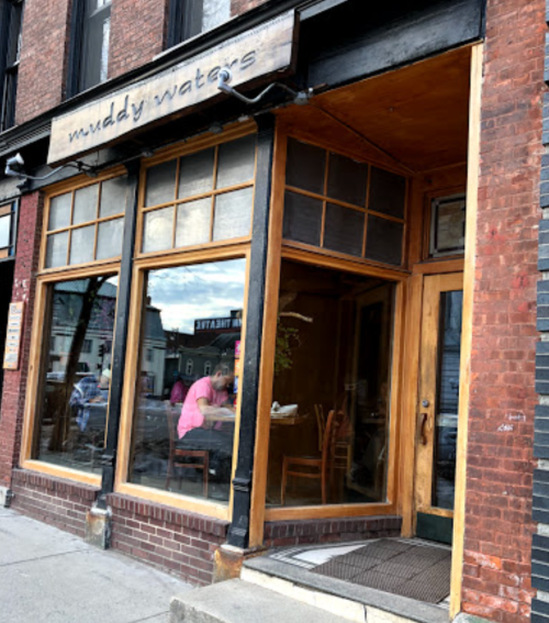 Exterior of Muddy Waters café, featuring large windows and a wooden door, with a person visible inside.