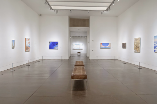A spacious art gallery with white walls, featuring various paintings and a brown bench in the center.