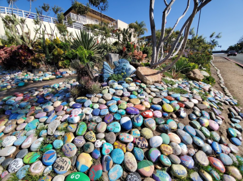 A colorful display of painted rocks in a landscaped area, surrounded by plants and trees under a clear blue sky.