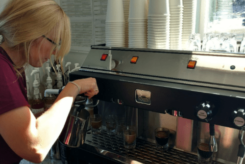 A barista prepares espresso shots using an espresso machine in a bright café setting.