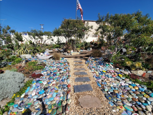 A colorful stone pathway leads through a vibrant garden with various plants and an American flag in the background.