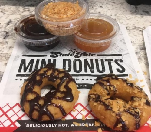 Two mini donuts topped with chocolate drizzle and coconut, accompanied by three dipping sauces on a branded paper.