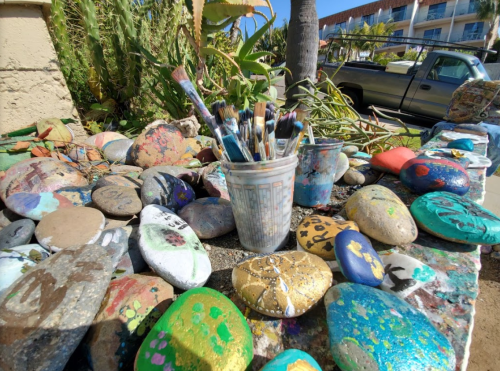 A collection of painted rocks and brushes in a cup, surrounded by greenery and a parked truck in the background.