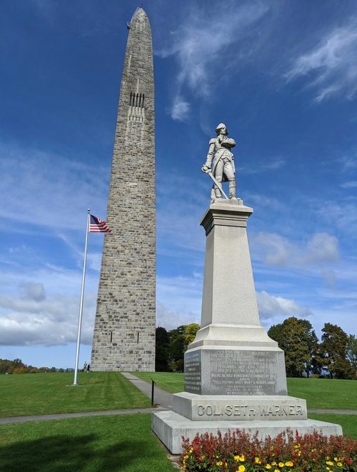 A tall stone monument with a statue of a soldier in front, set against a blue sky and green landscape.