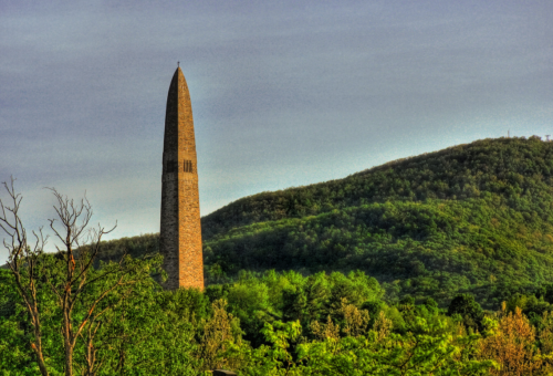 A tall stone monument stands amidst lush green trees, with a mountainous backdrop under a clear sky.