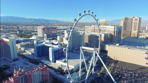 Aerial view of a large Ferris wheel in a cityscape with mountains in the background and various buildings nearby.