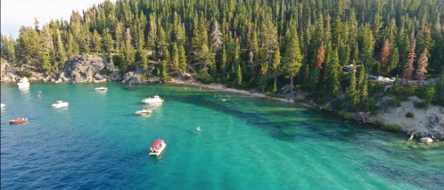 Aerial view of a clear blue lake surrounded by lush green trees, with boats scattered on the water.