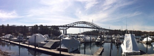 A panoramic view of a marina with covered boats and a large bridge in the background under a clear blue sky.