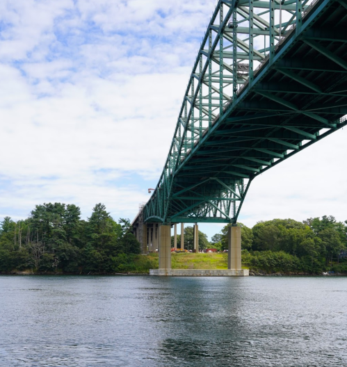 A green bridge spans over a calm river, surrounded by trees and a cloudy sky.