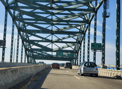 View of a highway under a large green bridge structure, with signs indicating the New Hampshire state line ahead.