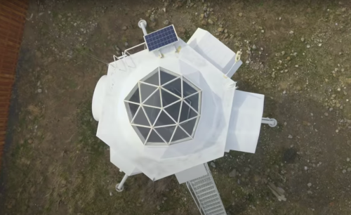 Aerial view of a geodesic dome structure with solar panels, surrounded by grass and rocks.