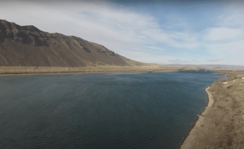 A serene river flows between mountains under a clear blue sky, with a sandy shore visible on the right.