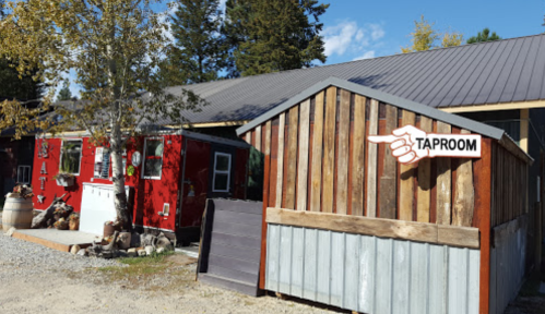 A rustic building with a sign pointing to a taproom, surrounded by trees and gravel.