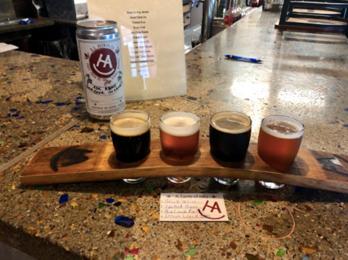 A wooden flight board with four beer samples and a can of beer on a bar counter.