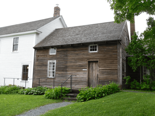 A rustic wooden house with a sloped roof, surrounded by greenery and a gravel path, next to a white building.
