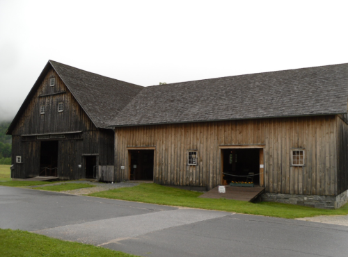 A large, rustic wooden barn with a sloped roof, set against a cloudy sky and green grass.
