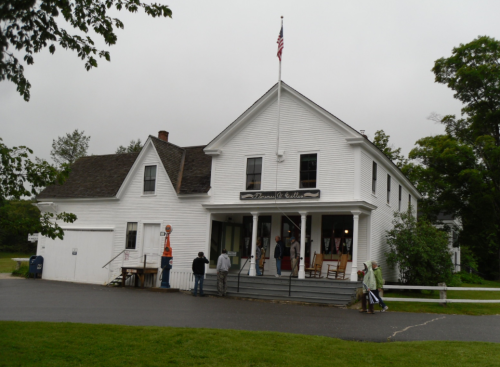 A white historic building with a porch, flag, and people gathered outside on a cloudy day.