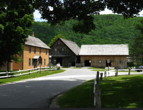 A scenic view of a rural area with two historic buildings and a tree-lined road under a blue sky.