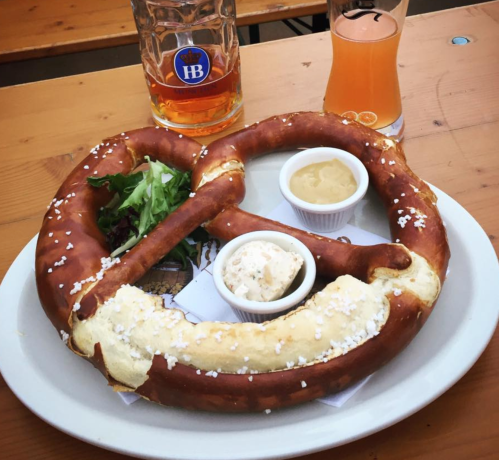 A large pretzel served with mustard and cheese, alongside a salad and two glasses of beer on a wooden table.
