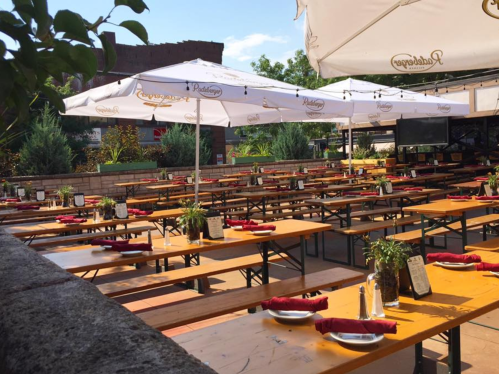 Outdoor dining area with wooden tables, benches, and umbrellas, set for guests with red napkins and potted plants.