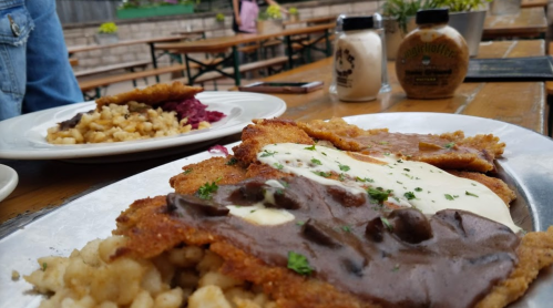 A plate of breaded schnitzel with gravy and creamy sauce, served with a side of pasta and a second dish in the background.