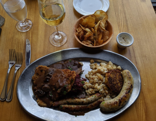 A metal platter with fried chicken, sausage, cauliflower, and gravy, accompanied by a bowl of chips and a glass of white wine.