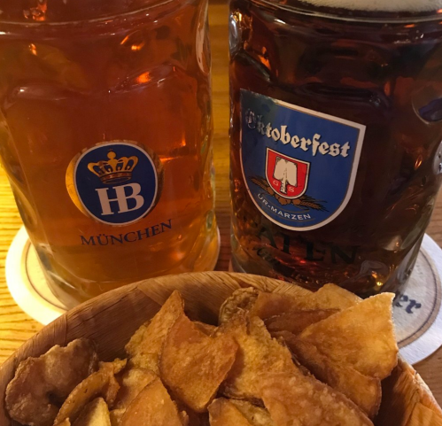 Two large beer mugs, one labeled "HB München" and the other "Oktoberfest," beside a bowl of crispy potato chips.