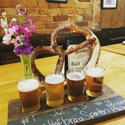 A large pretzel with a flower vase and a flight of four beer samples on a wooden board in a rustic setting.