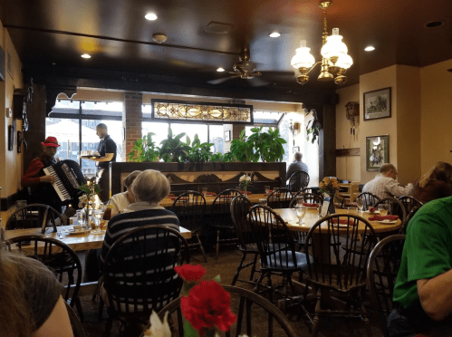 A cozy restaurant interior with plants, tables set for dining, and a musician playing accordion in the corner.