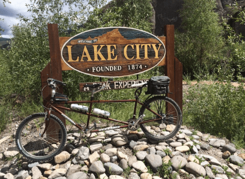 A tandem bicycle parked beside a wooden sign welcoming visitors to Lake City, established in 1874, surrounded by rocks.